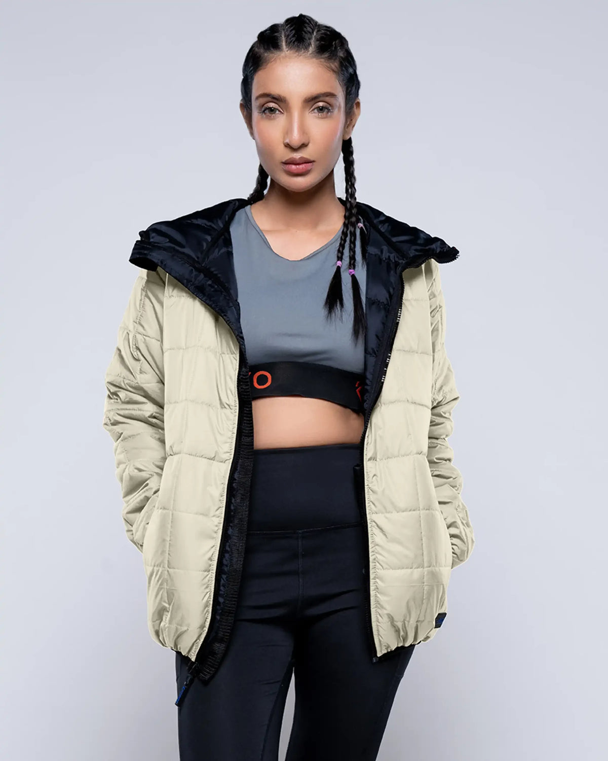 Woman wearing a beige puffer jacket with black interior, gray sports bra, and black leggings on a light gray background.