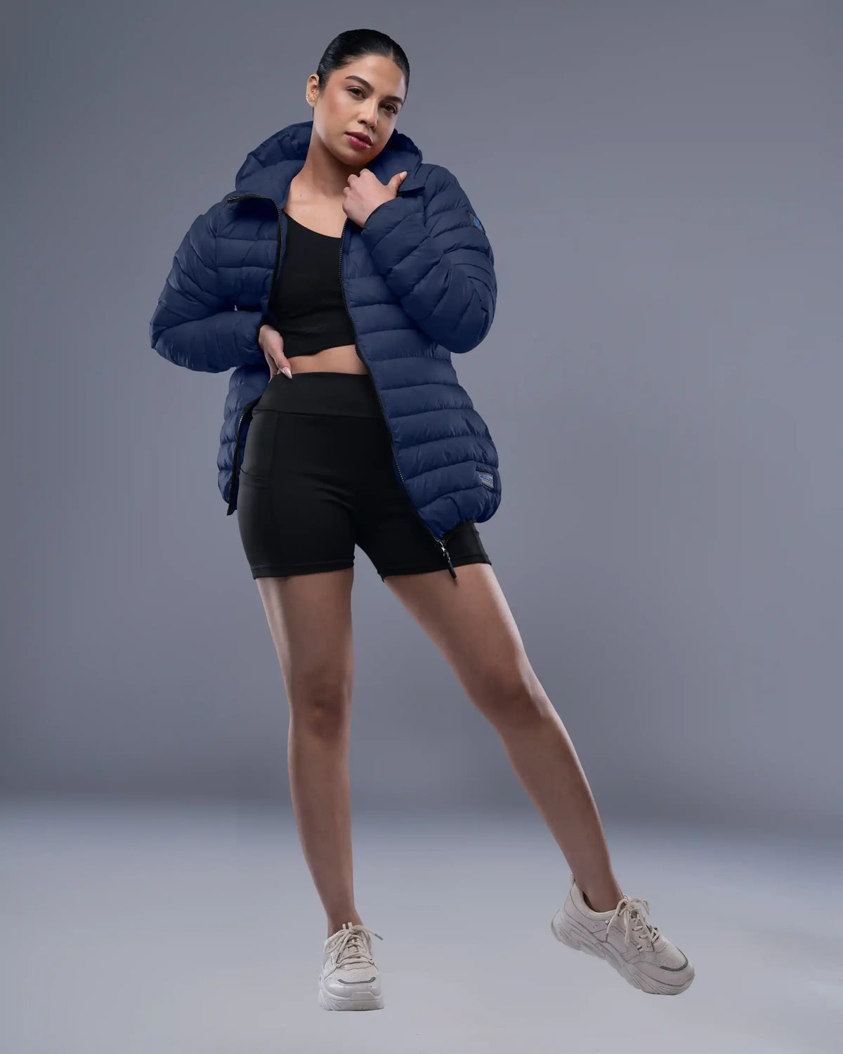 Woman wearing a blue puffer jacket, black athletic top, and shorts on a gray background