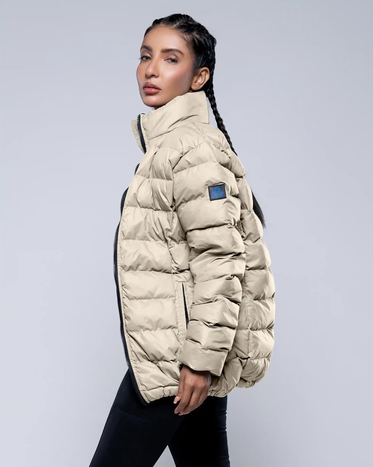 Woman wearing a beige puffer jacket with a blue patch on a light gray background