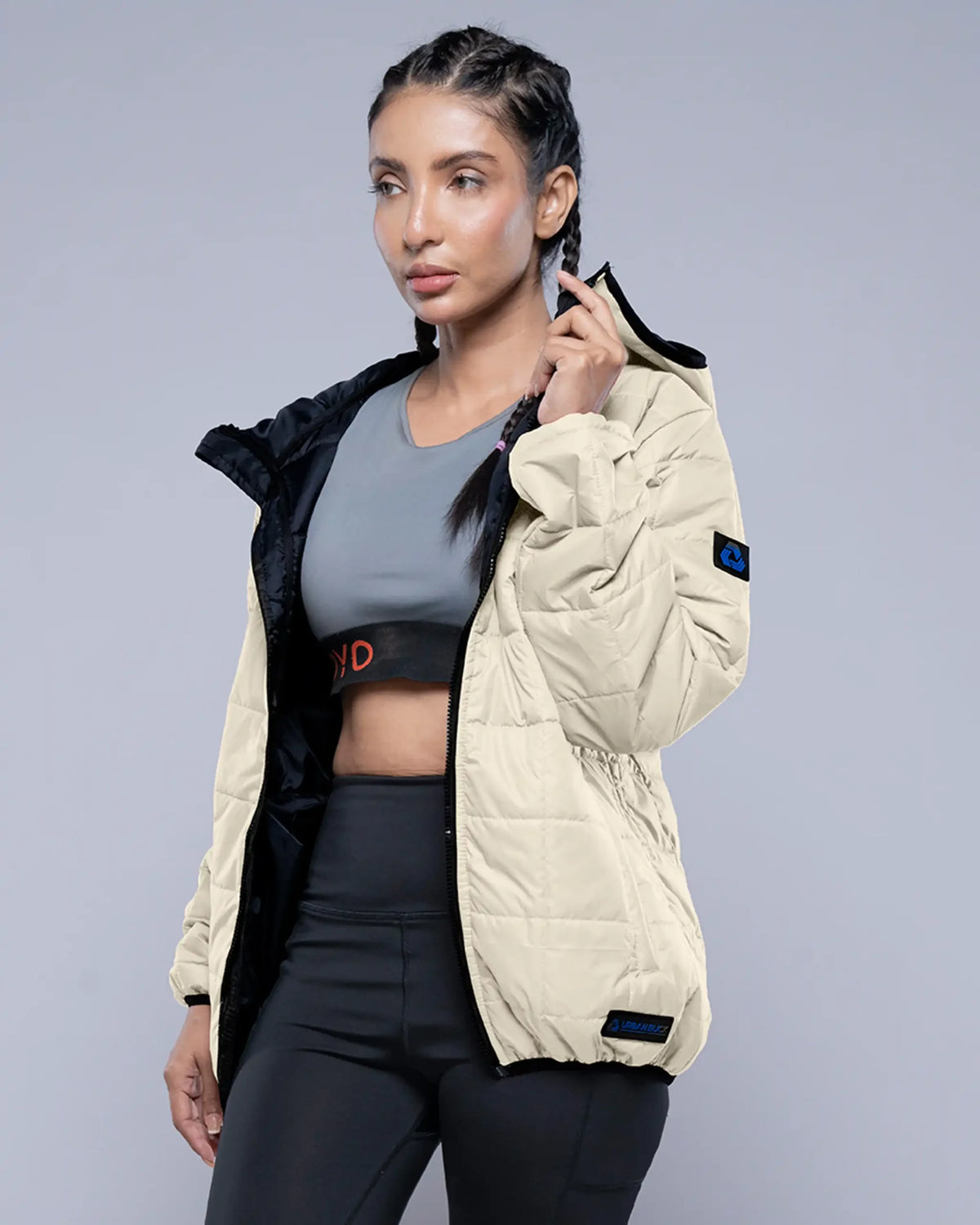 Woman wearing a beige quilted jacket over a gray sports bra and black leggings on a gray background