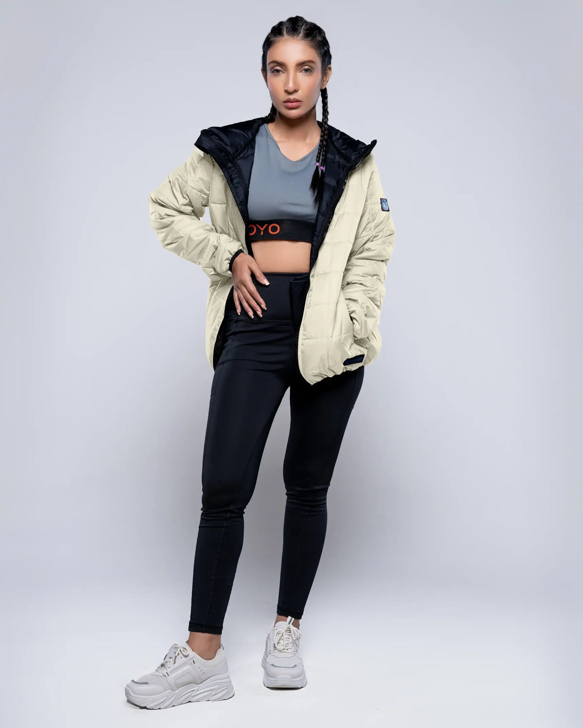 Woman wearing a beige puffer jacket, black leggings, and white sneakers on a light gray background