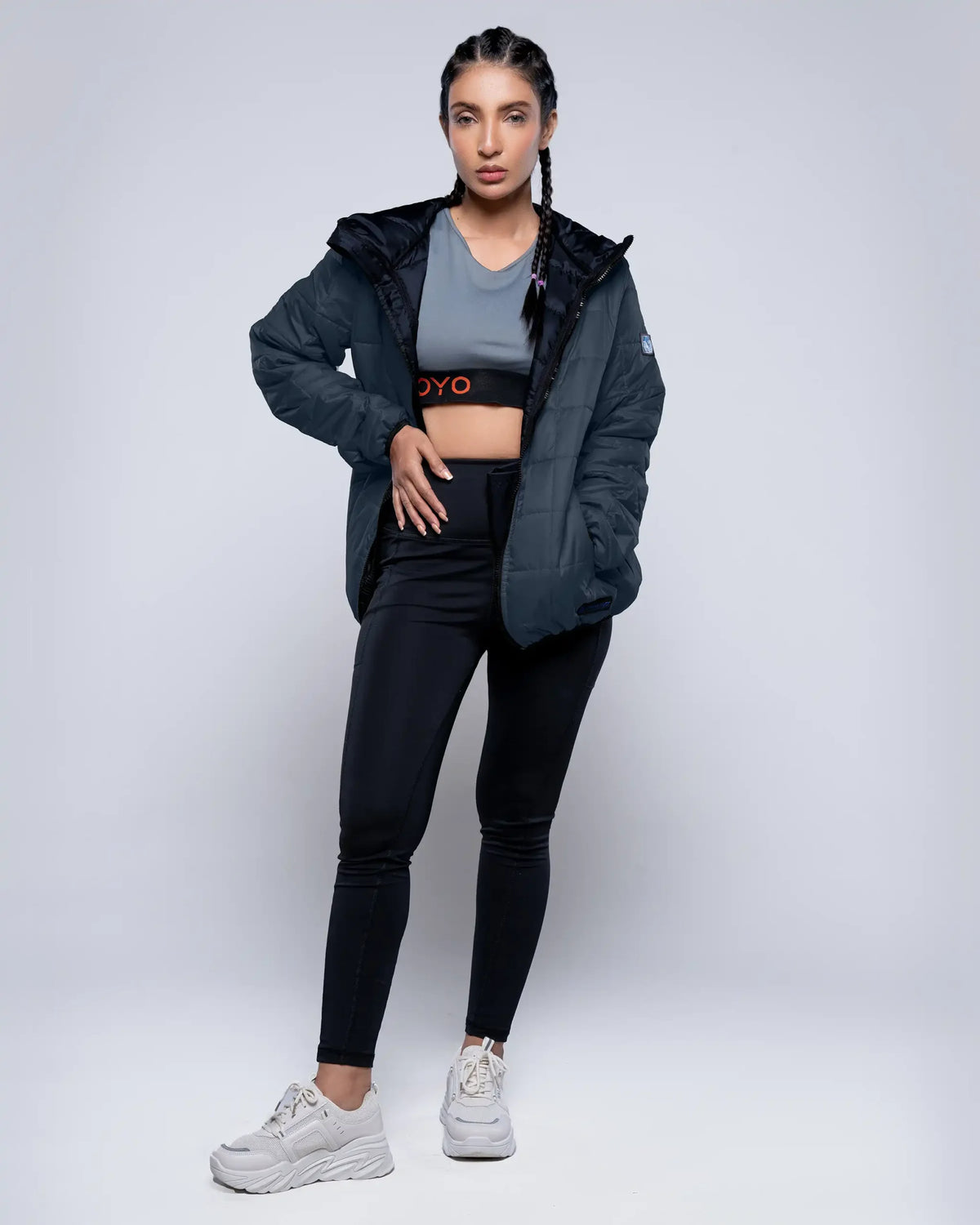 Woman wearing a black jacket, gray sports bra, black leggings, and white sneakers on a white background