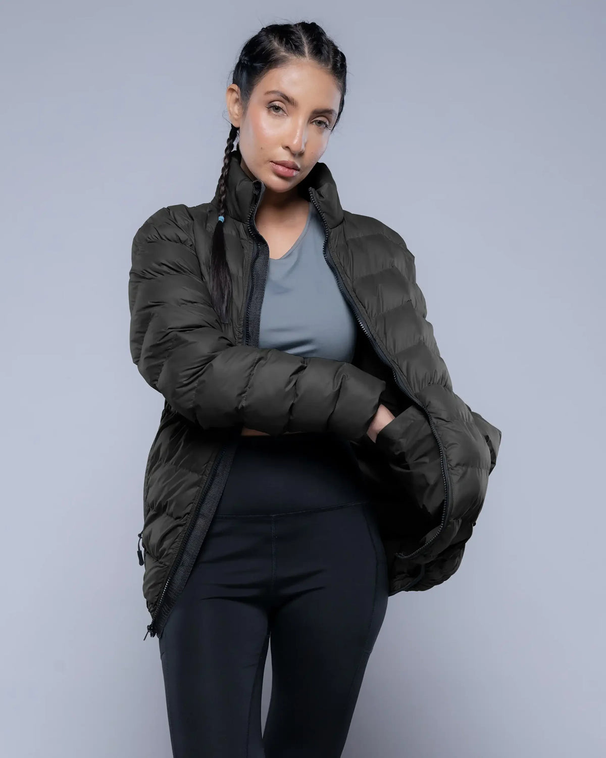Woman wearing a dark green puffer jacket over a gray top and black leggings on a light gray background