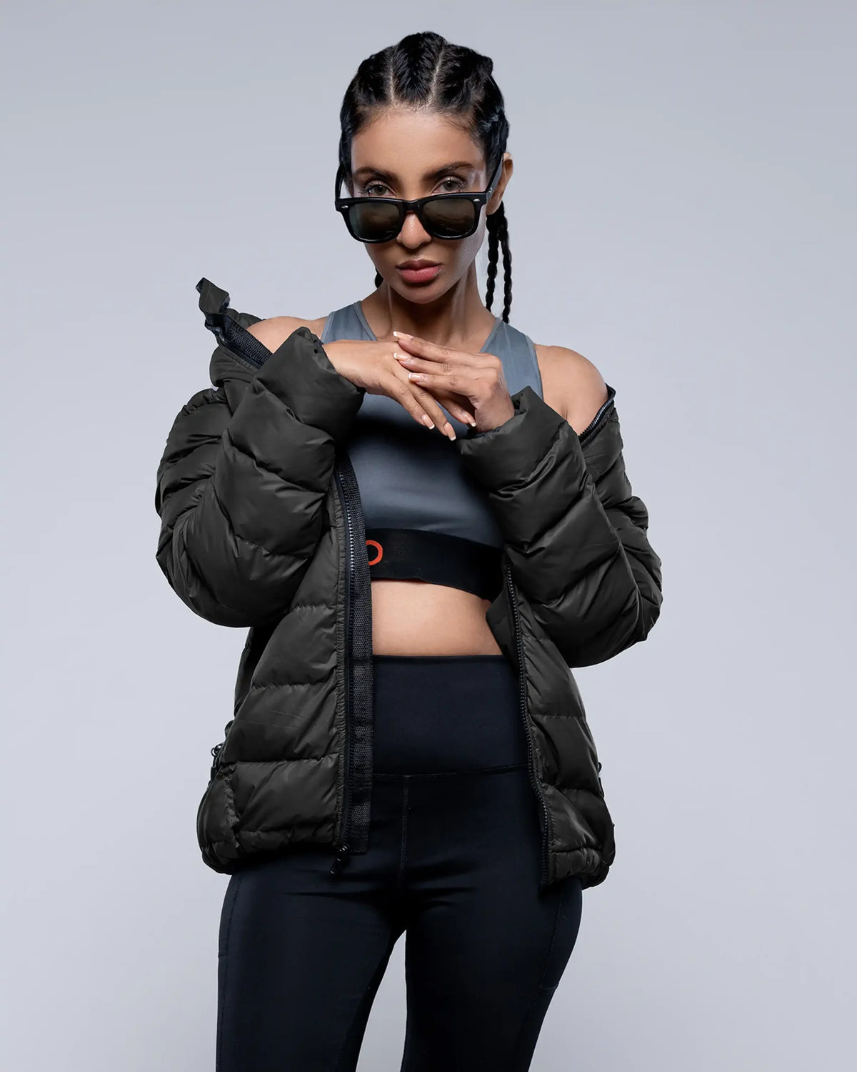 Woman wearing a black puffer jacket, sports bra, and leggings on a gray background