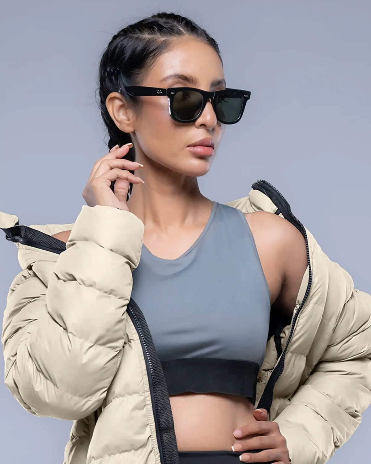 Woman wearing a beige puffer jacket, gray sports bra, and sunglasses against a gray background