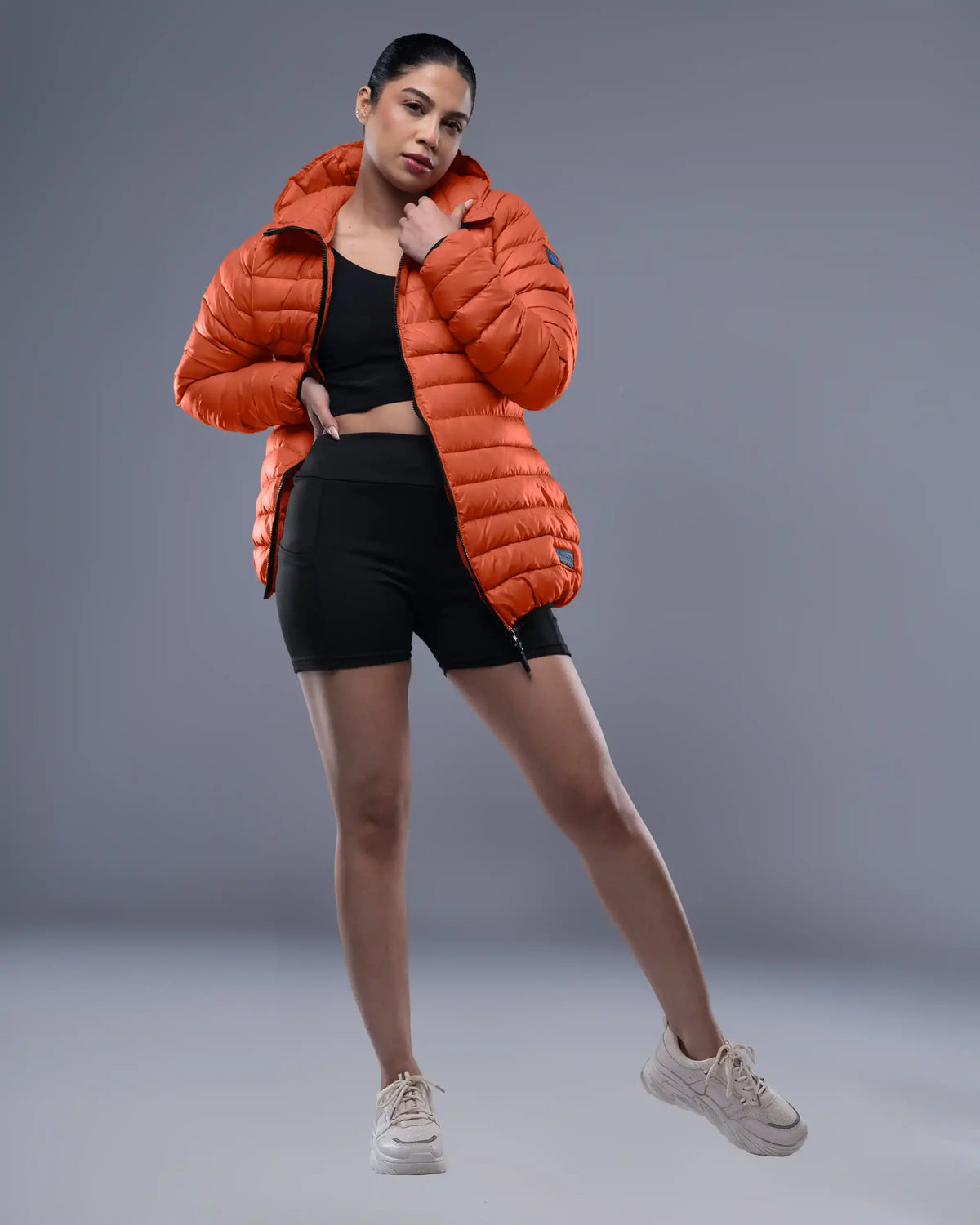 Person wearing an orange puffer jacket, black top, and shorts on a gray background